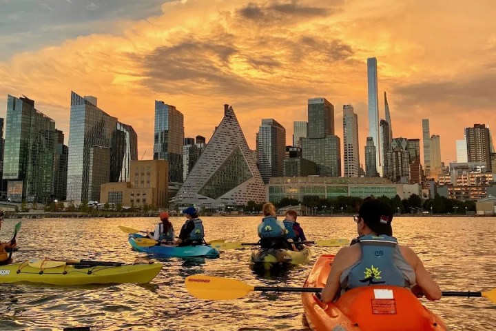 Kayakers in sit-on-top boats paddle on the Hudson River during a Skyline Kayak Trip, with a vibrant orange sunset behind the Midtown Manhattan skyline and the pyramid-shaped VIA building.
