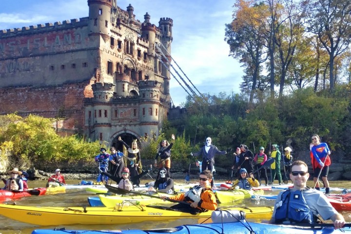A large group of Manhattan Kayak Co members in diverse Halloween costumes—including Superman, a pharaoh, and a skeleton—pose in their kayaks and paddleboards on the Hudson River with the historic ruins of Bannerman Castle in the background.