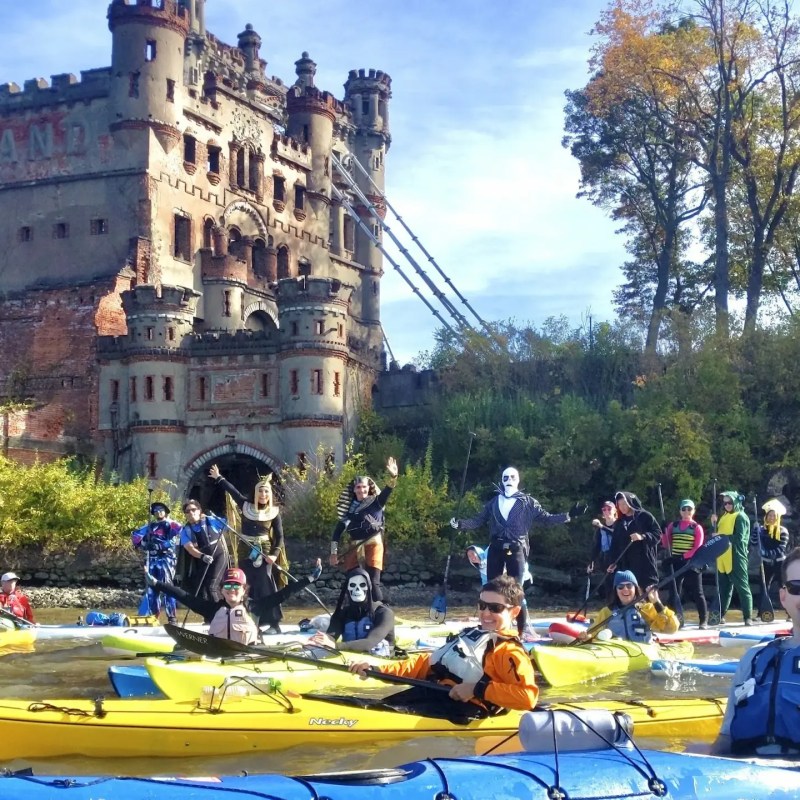 A large group of Manhattan Kayak Co members in diverse Halloween costumes—including Superman, a pharaoh, and a skeleton—pose in their kayaks and paddleboards on the Hudson River with the historic ruins of Bannerman Castle in the background.