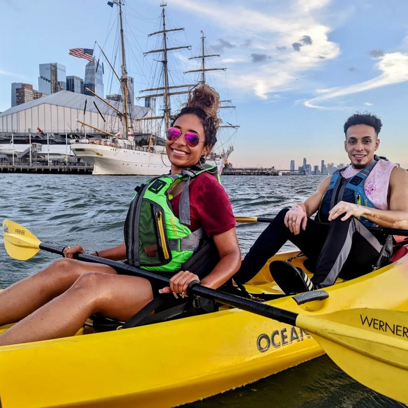Skyline Kayak Trip participants paddling a yellow sit-on-top kayak past the Mexican Navy training ship Cuauhtémoc and the USS Intrepid Museum at Pier 86.