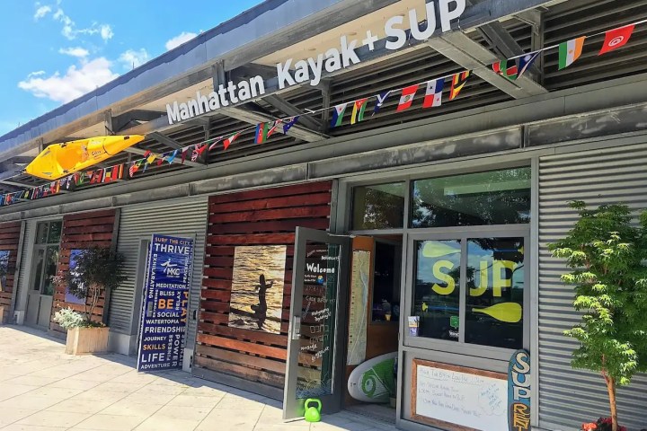 Front of Manhattan Kayak boathouse with colorful flags hung along the roofline and a yellow kayak displayed above the entry door.