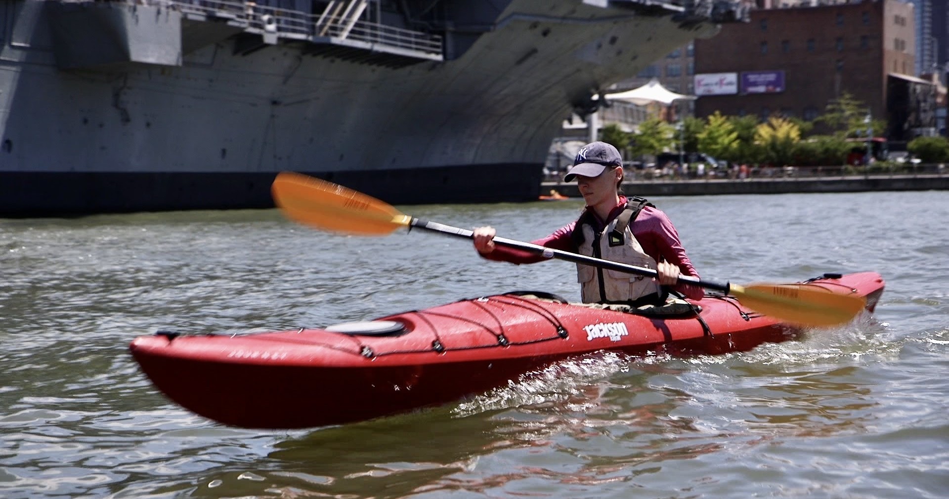 A focused student demonstrates the kinetic chain of a proper forward stroke in a red Jackson sea kayak. After a shore-based technical drill, she successfully applies her mechanics to the Hudson River during a private Manhattan Kayak Co lesson near the USS Intrepid.