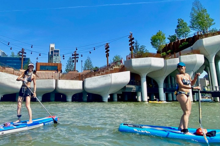 Two women on stand up paddle boards in front of Little Island on the Hudson River in NYC