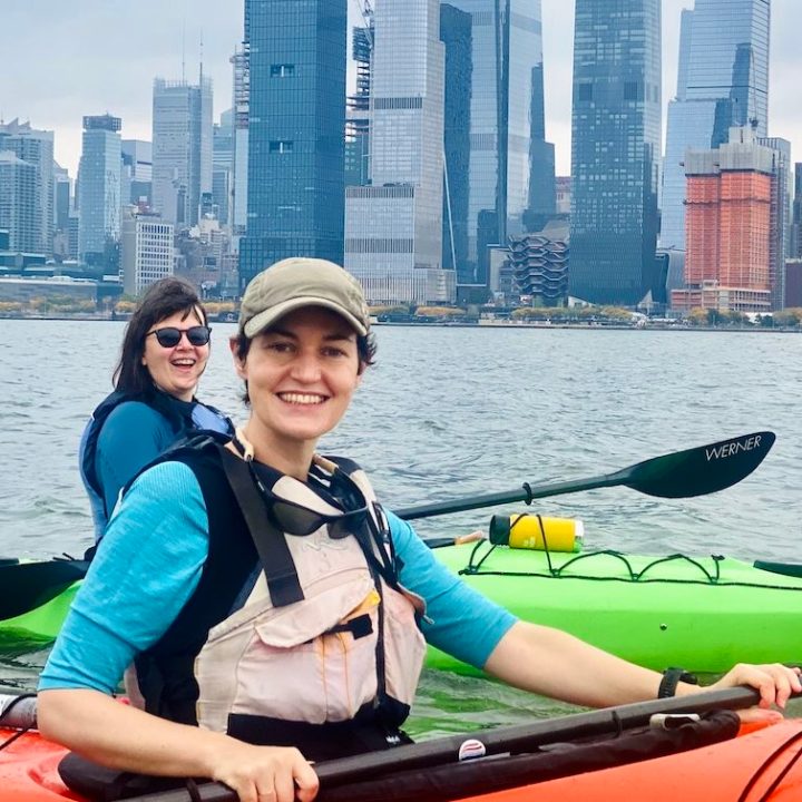 two smiling women in sea kayaks in front of the Hudson Yards on the Hudson River in NYC