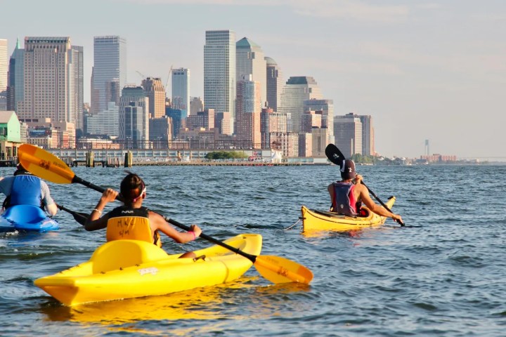 Professional guidance on the Hudson: A Manhattan Kayak guide leads two beginners in stable sit-on-top kayaks toward the distant downtown NYC skyline, demonstrating the safe and relaxed pace of our guided river tours.