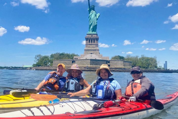 three women and a man in kayaks in front of the Statue of Liberty