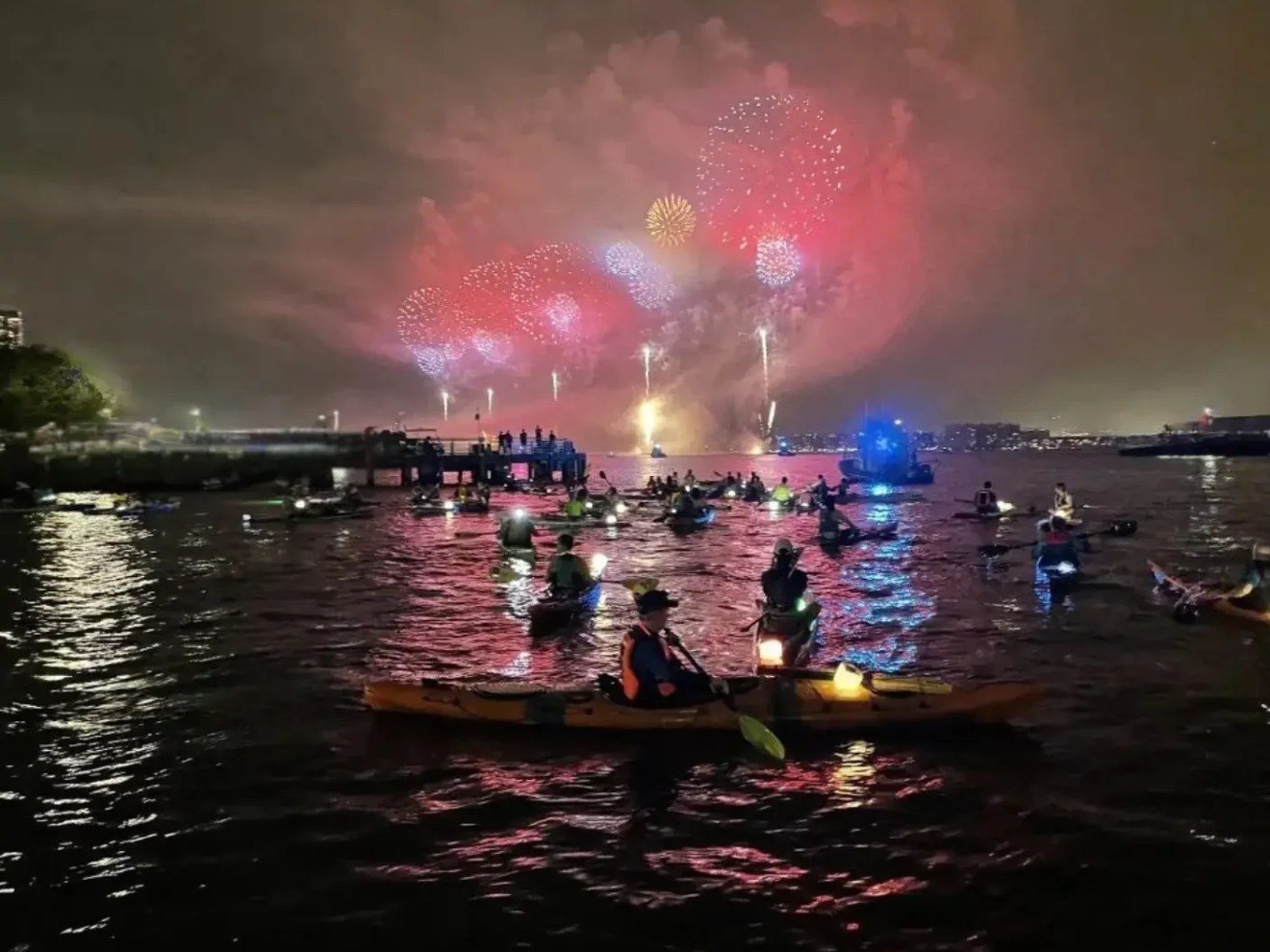 A fleet of Manhattan Kayak members in illuminated kayaks gather on the dark Hudson River, watching a spectacular display of red and gold July 4th fireworks exploding over the New York City skyline.