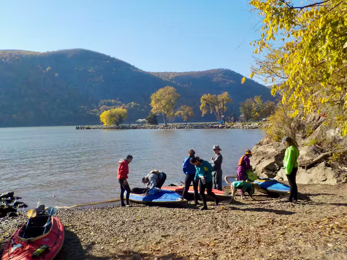 A group of kayakers along a riverfront beach surrounded by yellow foliage and hills. An exclusive day trip for Manhattan Kayak members during peak fall foliage in the Hudson Valley.