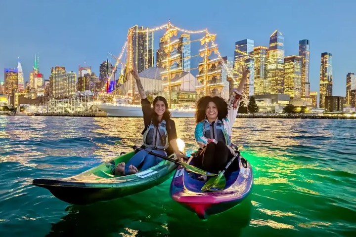 Two friends cheer with raised arms while night kayaking in the Hudson River. Their green LED-lit kayaks glow against a stunning backdrop of the illuminated Mexican Navy tall ship, Hudson Yards, and the glowing Midtown Manhattan skyline.