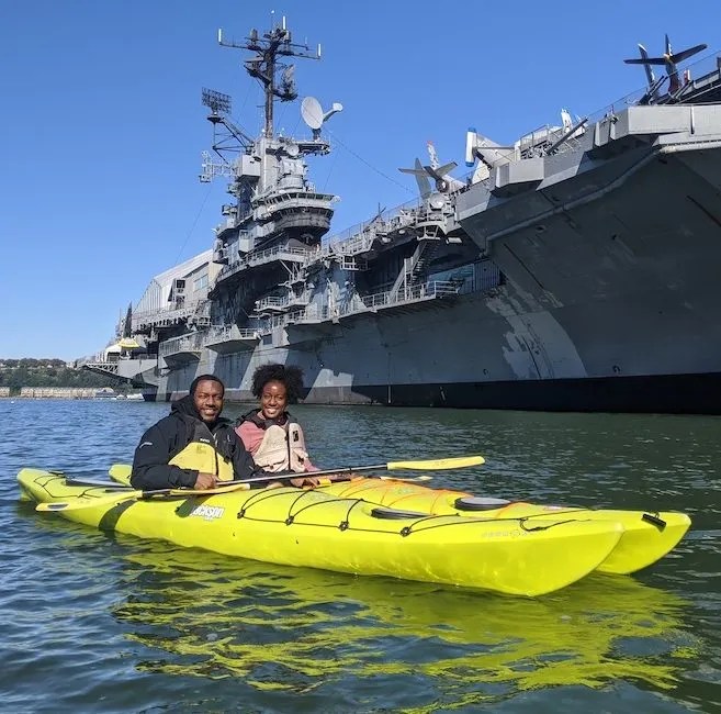 A happy couple enjoying a peaceful beginner kayak lesson in yellow touring kayaks, paddling past the historic Intrepid on the Hudson River.