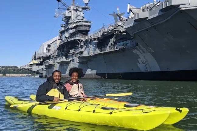 A happy couple enjoying a peaceful beginner kayak lesson in yellow touring kayaks, paddling past the historic Intrepid on the Hudson River.