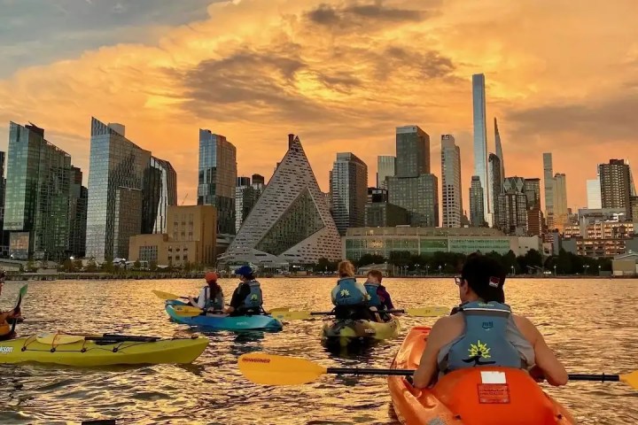 Kayakers in sit-on-top boats paddle on the Hudson River during a Skyline Kayak Trip, with a vibrant orange sunset behind the Midtown Manhattan skyline and the pyramid-shaped VIA building.