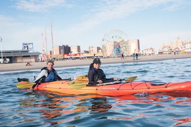 A couple in a tandem sea kayak near the beach with the Coney Island skyline in the background.