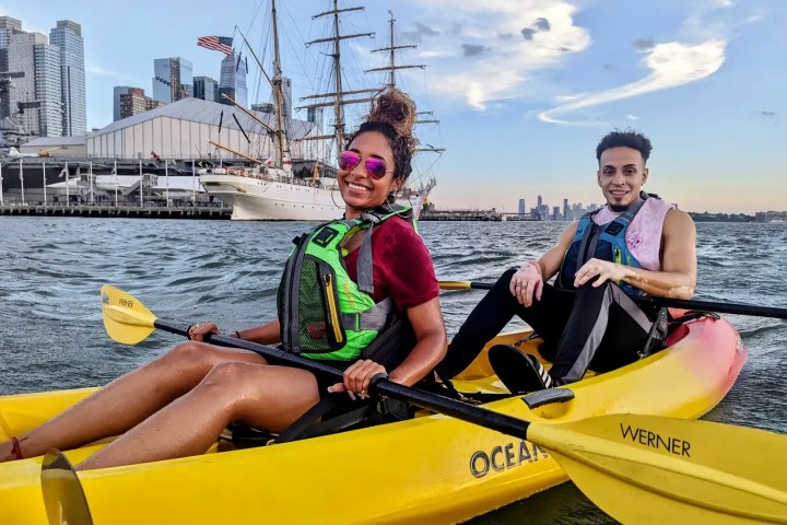Skyline Kayak Trip participants paddling a yellow sit-on-top kayak past the Mexican Navy training ship Cuauhtémoc and the USS Intrepid Museum at Pier 86.
