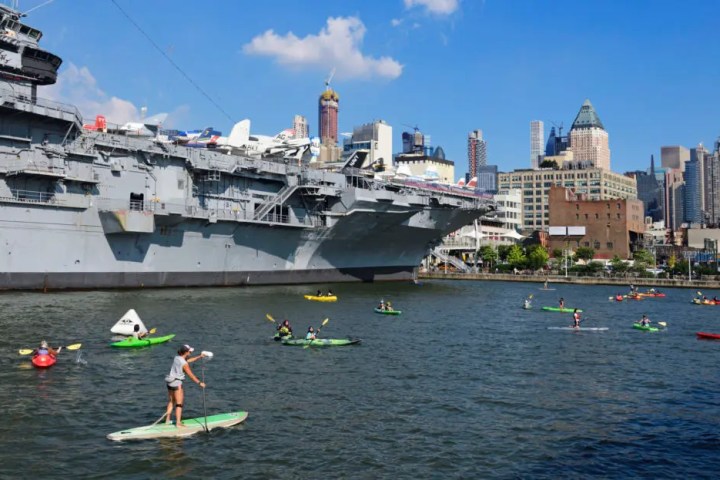 Kayakers and paddleboarders paddling near the Intrepid aircraft carrier with the Midtown Manhattan skyline in the background under a bright blue sky.