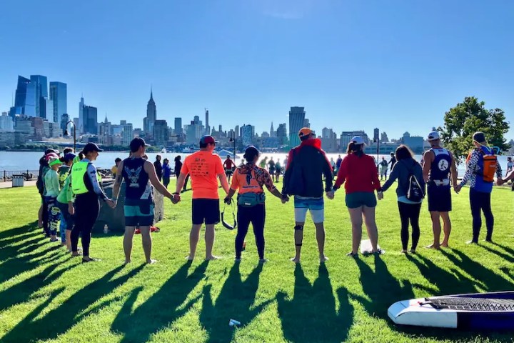 Manhattan Kayak Co Adventure Club members join a larger group of racers on the green grass of Maxwell Place Park in Hoboken, New Jersey. They stand in a circle holding hands with the morning sun casting their shadows behind them, before the Hudson River Cup race. The iconic Manhattan skyline stands tall across the sparkling river under a clear blue sky, framing the group in a moment of unity and anticipation.