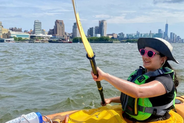 A smiling Kayak Basics student demonstrating a rhythmic core-driven stroke during her Class 3 graduation paddle on the Hudson River, with the Chelsea skyline in the distance.
