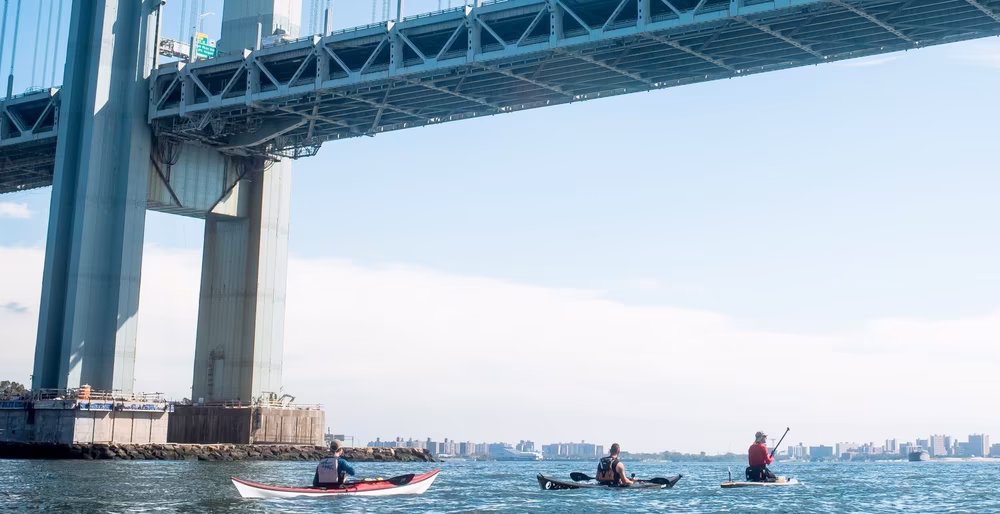 A group of Manhattan Kayak Co kayakers under the Verrazzano-Narrows Bridge on a blue summer day. The Lower NY Bay and Atlantic Ocean are on the horizon.