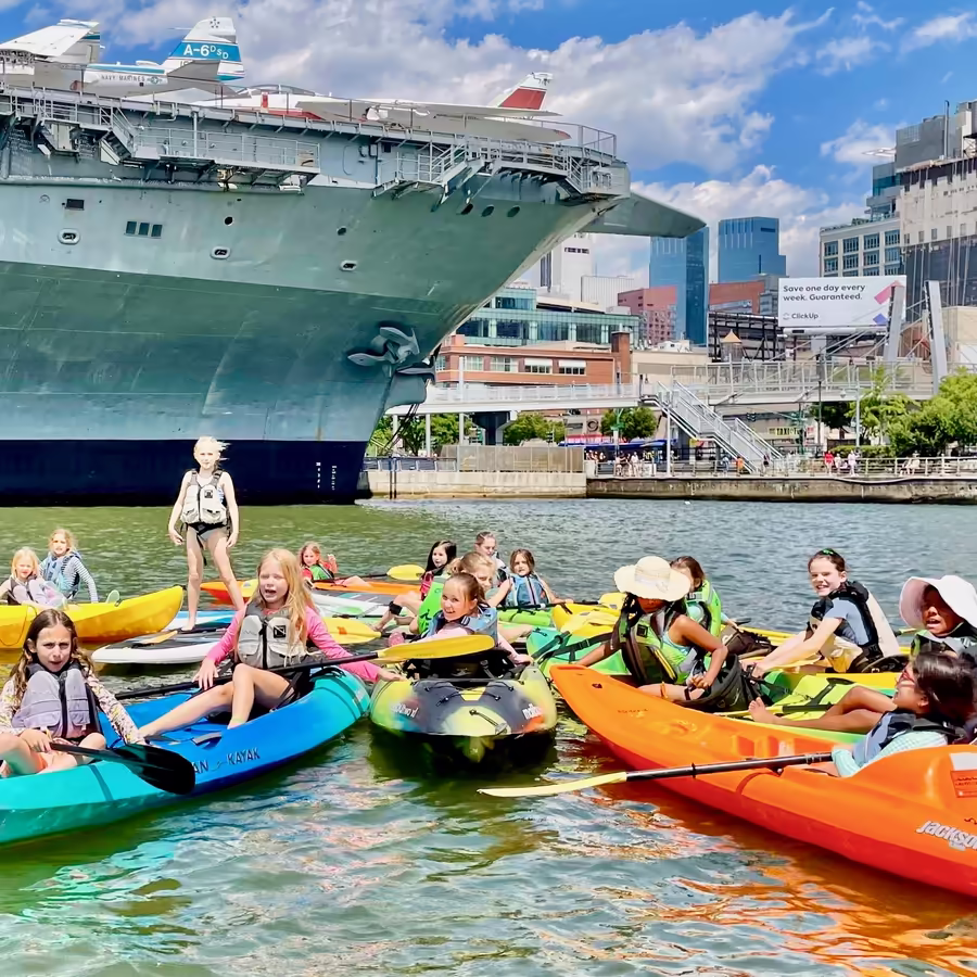 A large group of kids having a birthday party in the safe sanctuary cove at Manhattan Kayak. Popular NYC kids' birthday party idea at Pier 84, featuring kayaking and space for a picnic and cake in the park.