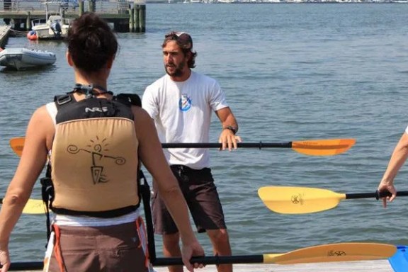 At the Pier 84 dock, a professional Manhattan Kayak instructor faces a student to demonstrate the correct paddle grip, with the calm Hudson River in the background. The student in the foreground wears a tan life jacket.