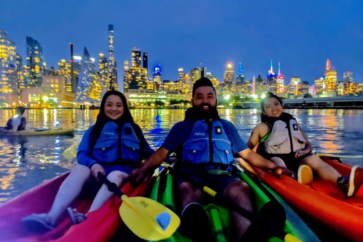 Three friends smiling in glowing LED-lit kayaks smile together on the Hudson River, with the iconic Midtown Manhattan skyline and the pyramid-shaped VIA 57 West building illuminated behind them during a night kayaking tour in NYC.