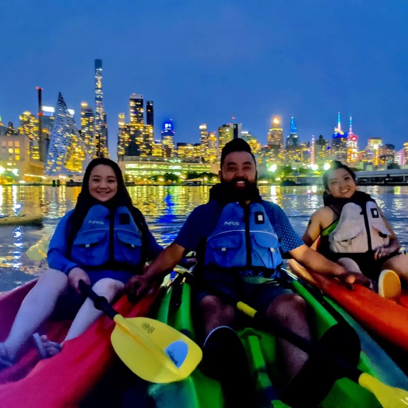 Three friends smiling in glowing LED-lit kayaks smile together on the Hudson River, with the iconic Midtown Manhattan skyline and the pyramid-shaped VIA 57 West building illuminated behind them during a night kayaking tour in NYC.