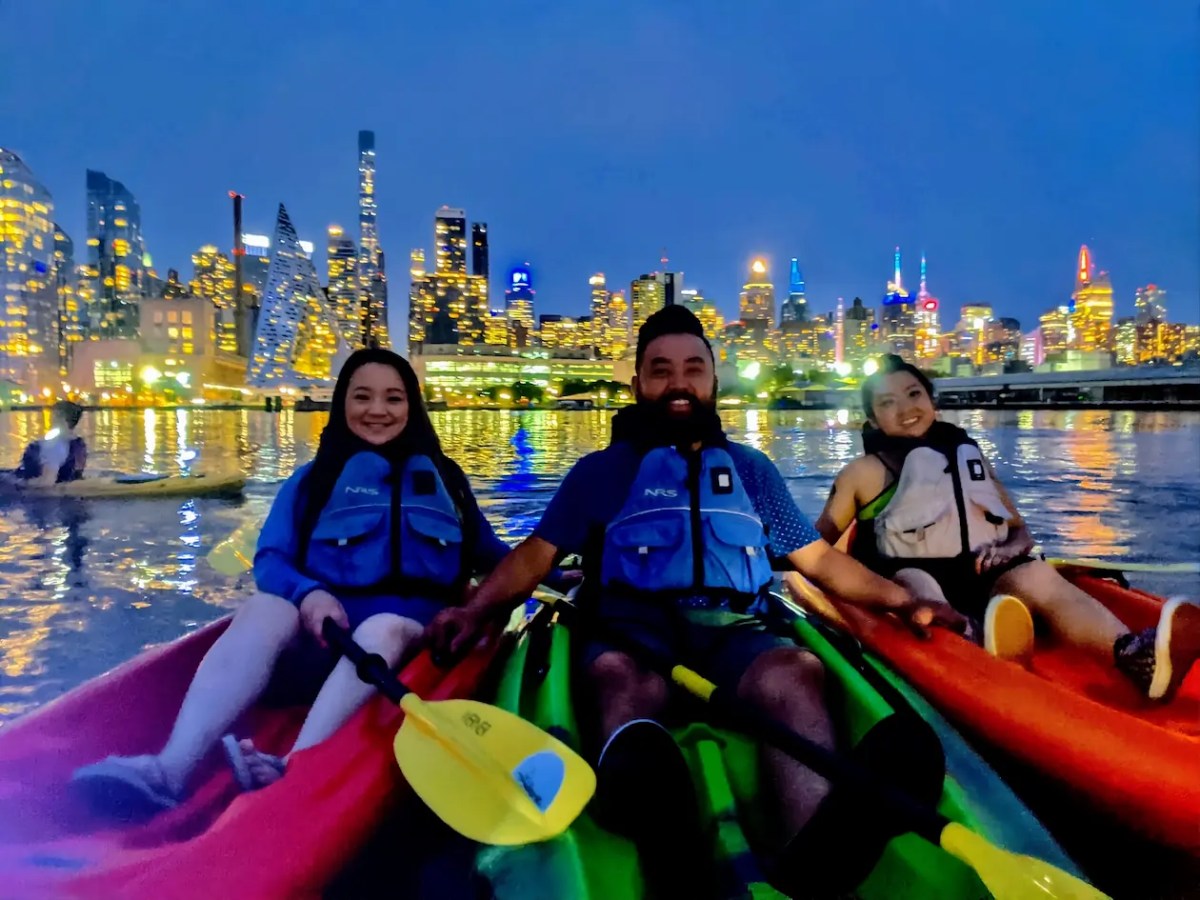Three friends in glowing LED-lit kayaks smile on the Hudson River, with the illuminated Midtown Manhattan skyline and Hudson Yards glowing behind them during a night kayak tour.