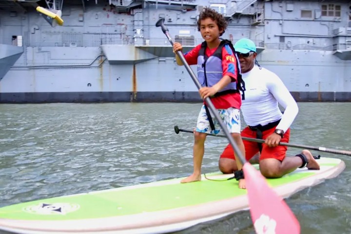 A young boy standing on a paddleboard with his father kneeling behind him during the Pier 84 public rental program at Manhattan Kayak Co.