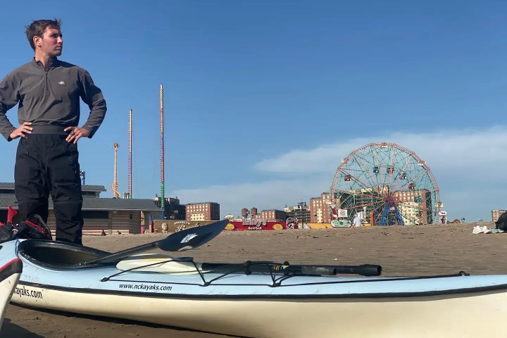 A man wearing outdoor gear stands along his sea kayak on Coney Island Beach & Boardwalk with a blue sky and Luna Park rides in the background.