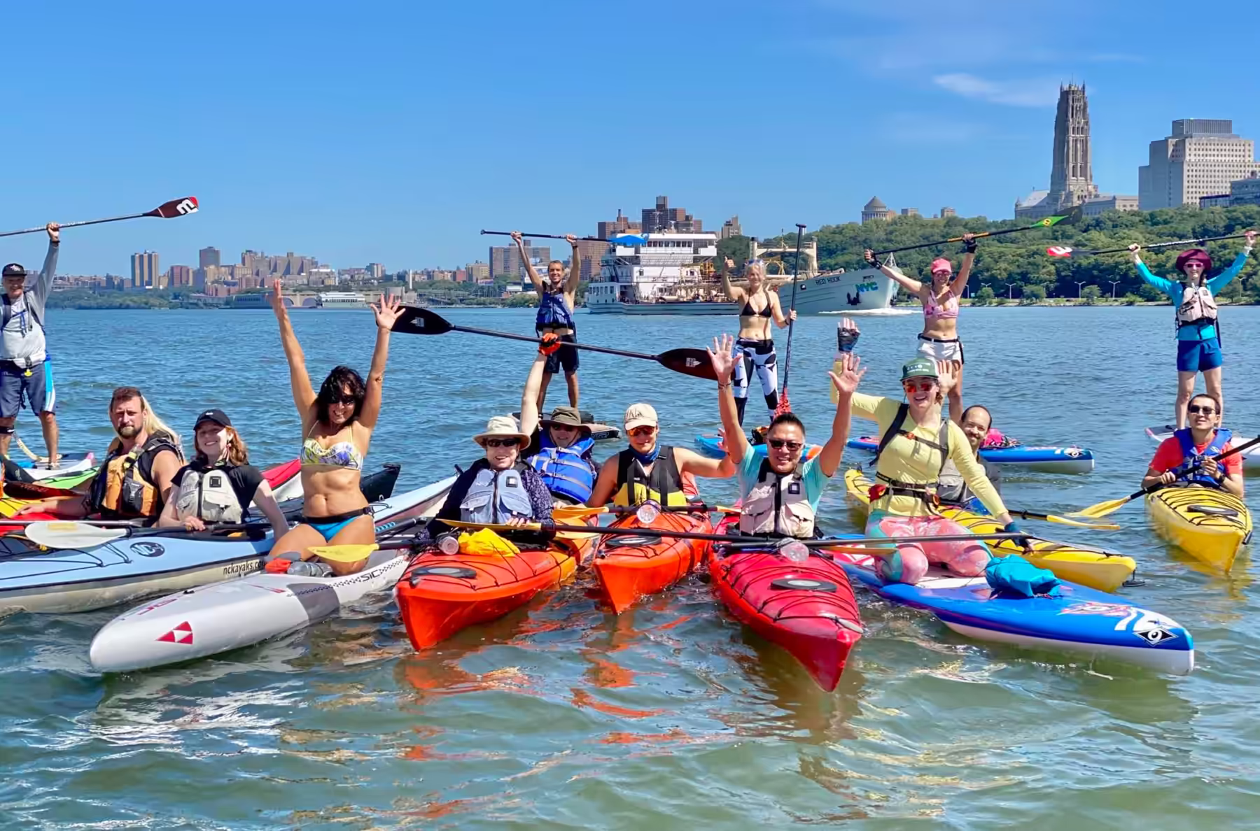A group of 15 Manhattan Kayak Co members on sea kayaks and paddleboards on the Hudson River, raising their paddles and laughing under a blue sky.