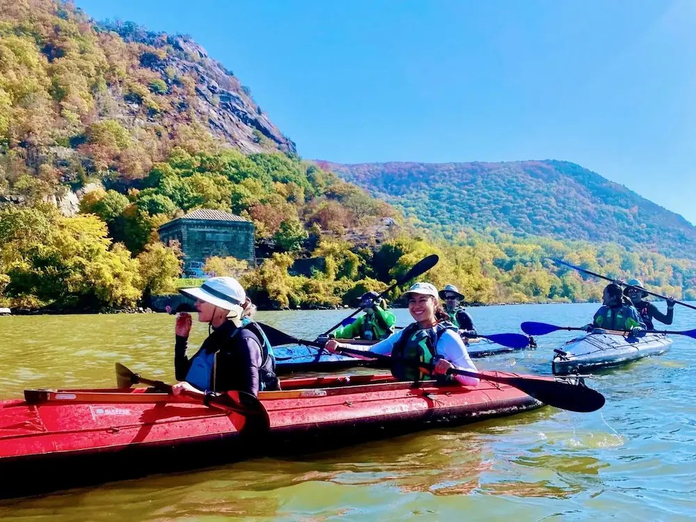 A joyful group of six Manhattan Kayak Co Adventure Club members paddle tandem sea kayaks past the Breakneck Tunnel, enjoying the peak fall foliage of the Hudson Highlands during an exclusive members-only day trip from NYC.