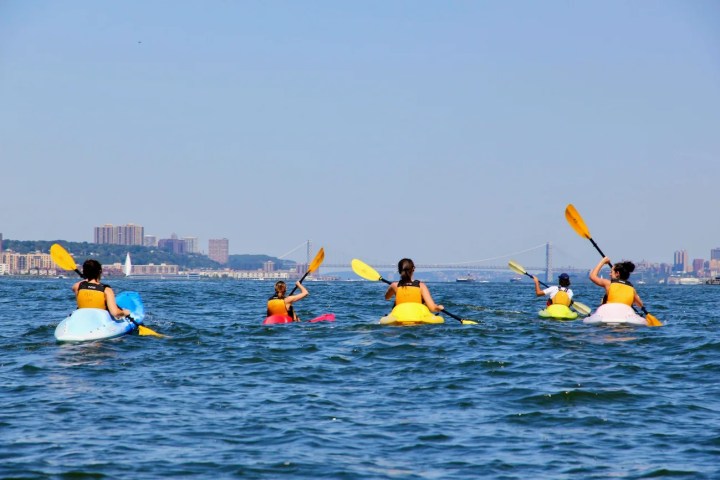 Heading north into the open blue: Five paddlers in stable, colorful sit-on-top kayaks navigate the wide expanse of the Hudson River toward the distant George Washington Bridge, with Harlem and Fort Lee visible on the horizon under a clear sky.