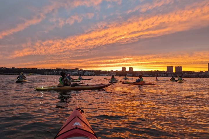 A serene moment on the Hudson River: Beginner kayakers pause to relax and take photos of a vivid orange sunset over Weehawken, New Jersey, with the fiery sky reflecting beautifully on the calm water.