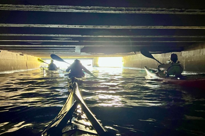 An gritty, industrial POV from a sea kayak paddling underground. Four club members navigate the dark, concrete underbelly of the city toward a bright light at the tunnel's exit.