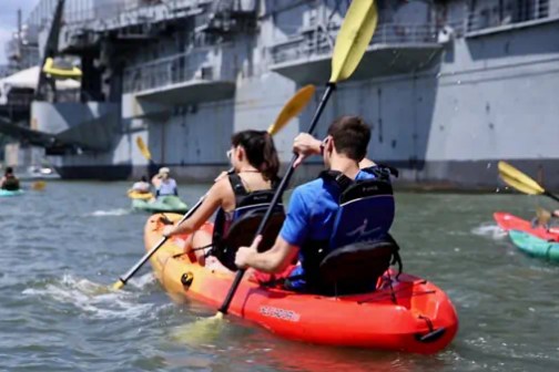 A couple riding a tandem sit-on-top kayak in front of the Intrepid Aircraft Carrier on the Hudson River in NYC.