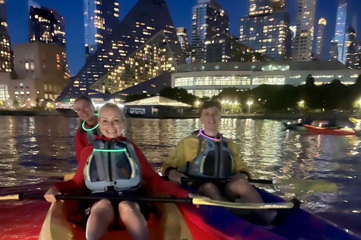 A group of friends laughing in glowing, LED-lit kayaks on the Hudson River at night. They wear neon glow necklaces with the lit-up Midtown Manhattan skyline and the iconic VIA 57 West pyramid building as their backdrop—the ultimate alcohol-free summer nightlife experience in NYC.