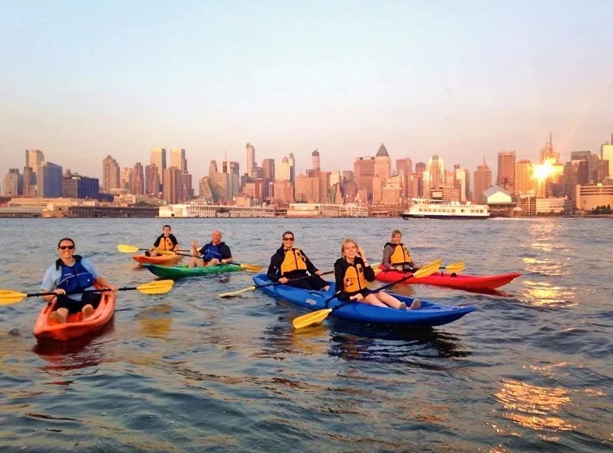 Wide angle sunset photo of 6 people riding sit-on-top kayaks on the Hudson River with the Midtown skyline behind them.