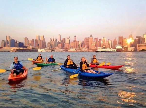 Wide angle sunset photo of 6 people riding sit-on-top kayaks on the Hudson River with the Midtown skyline behind them.