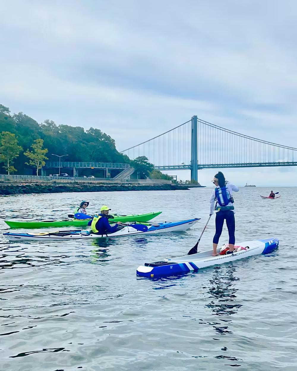 A woman riding an SIC Atlantis paddleboard and three sea kayakers approaching Verrazzano Bridge.