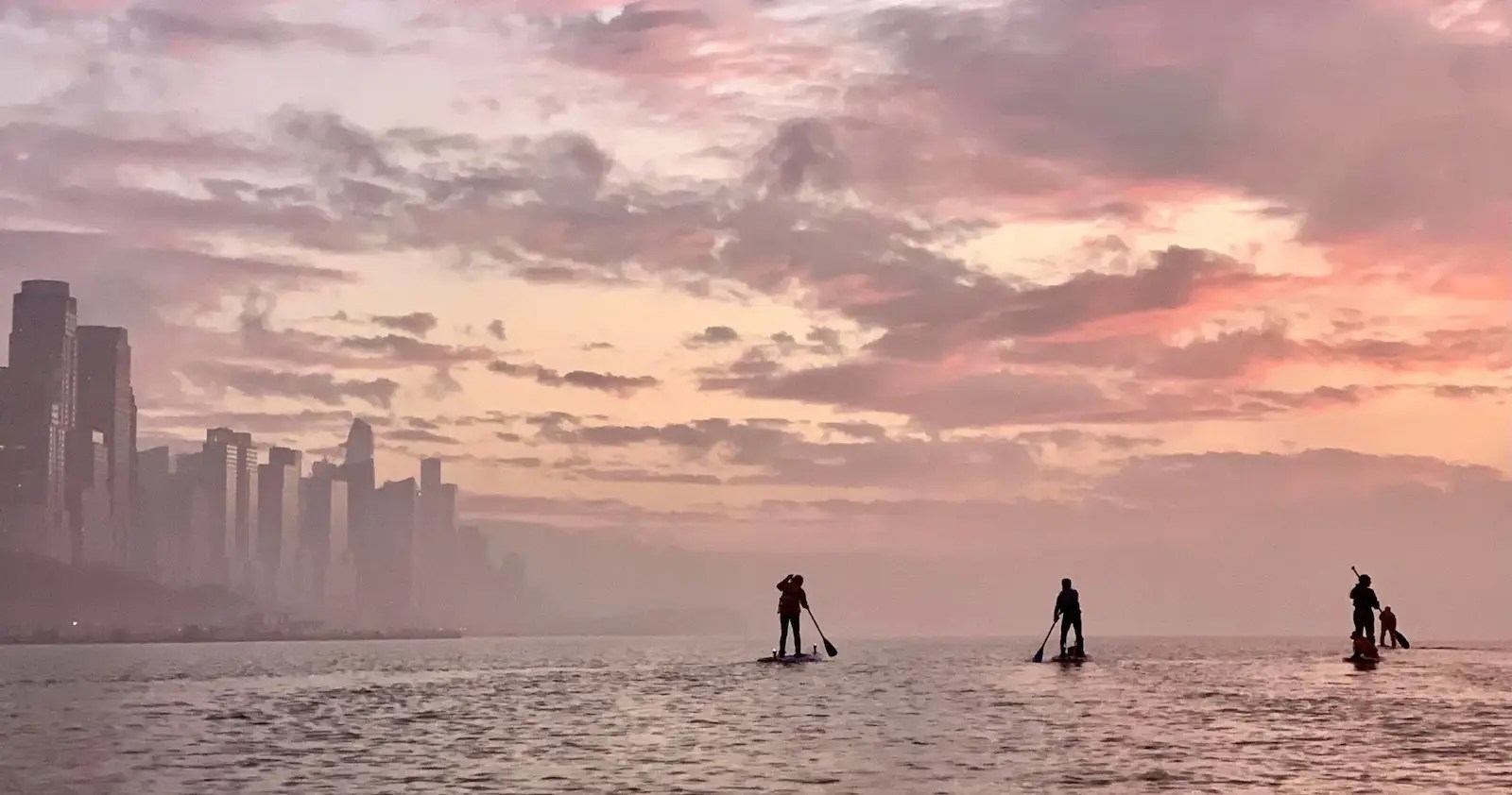Silhouetted paddleboarders on the Hudson River at sunset with the Upper West Side skyline on their left and an open expanse all around..