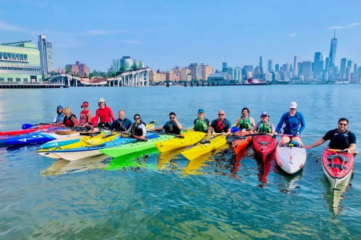 Thirteen smiling kayakers and paddleboarders rafted alongside each other on the Hudson River with Pier 57, Little Island, One World Trade, and a clear blue sky in the background.