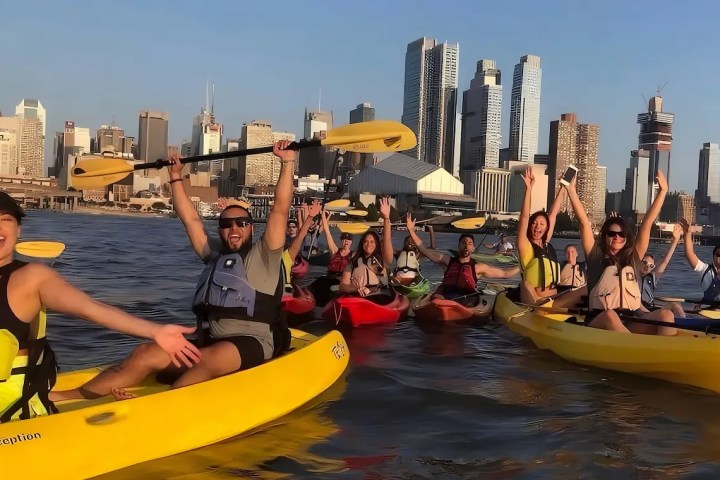 A large, enthusiastic corporate team cheering and raising paddles during a private Hudson River kayak trip with the Midtown Manhattan skyline in the background.