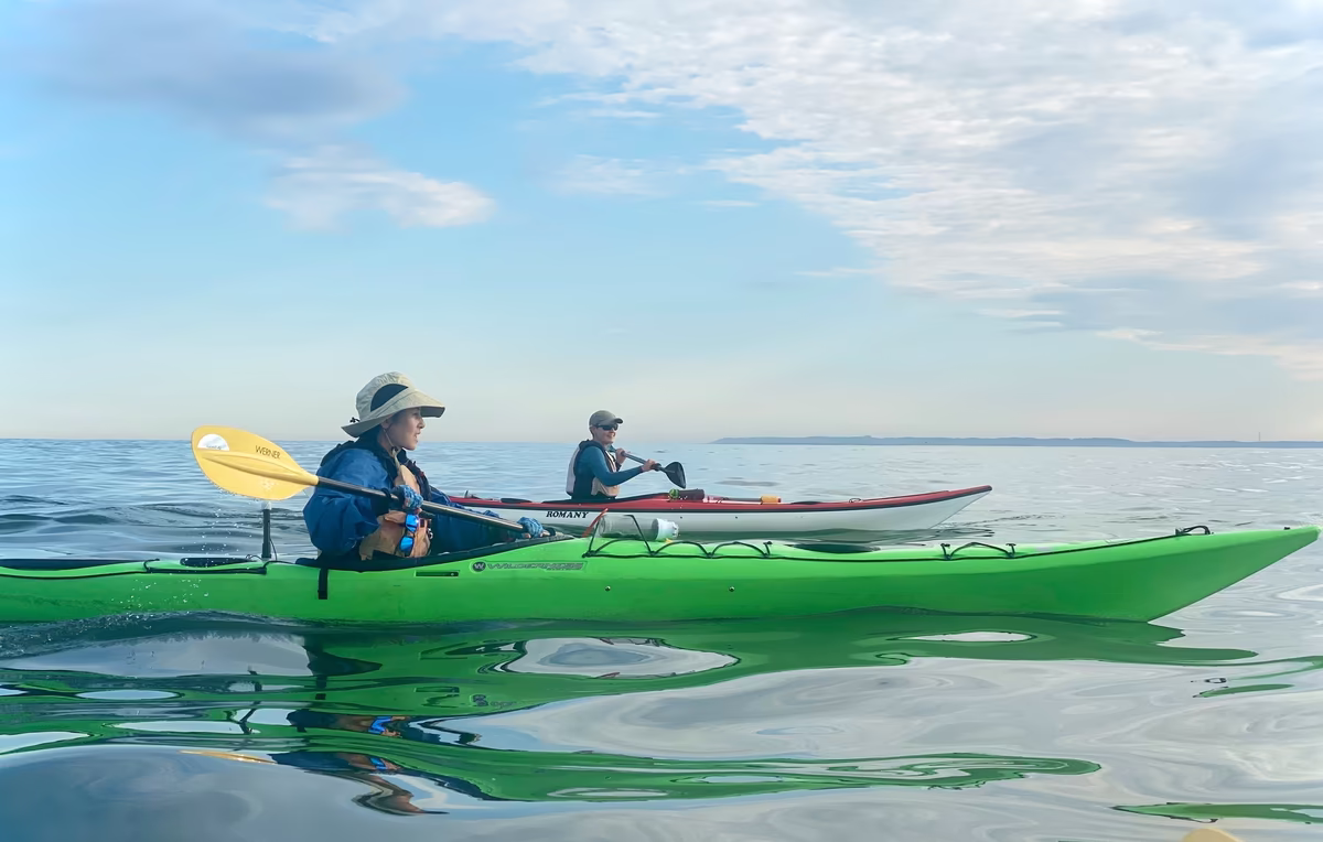 Two women riding sea kayaks–a lime green Wilderness Systems Tempest 170 and a red and white NDK Romany. They are kayaking in the Lower NY Harbor on calm reflective water with a completely open horizon and partly cloudy sky beyond.