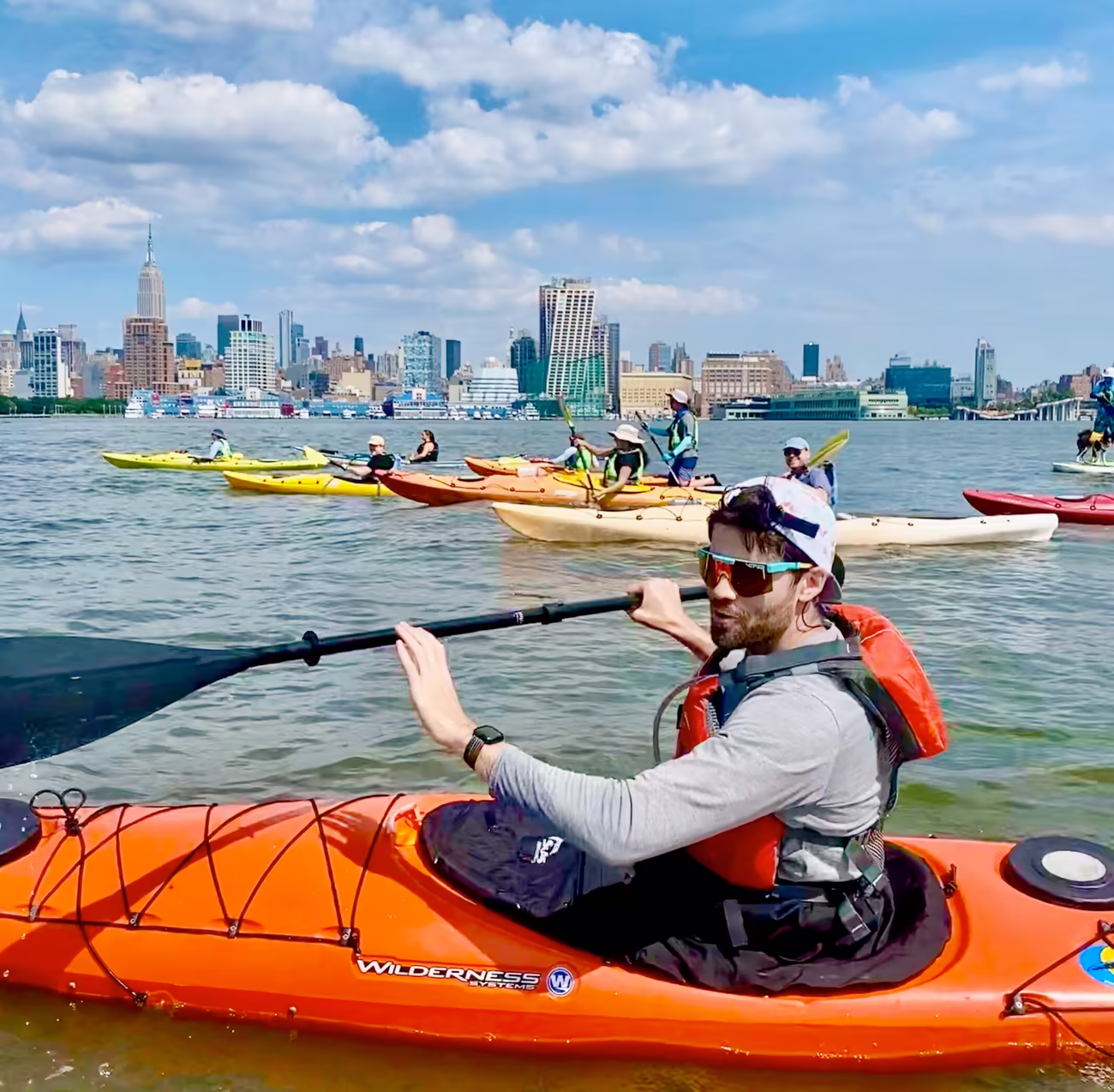 A man wearing sunglasses and riding an orange Wilderness Systems kayak on the Hudson River with group of paddlers and the Manhattan skyline in the background.
