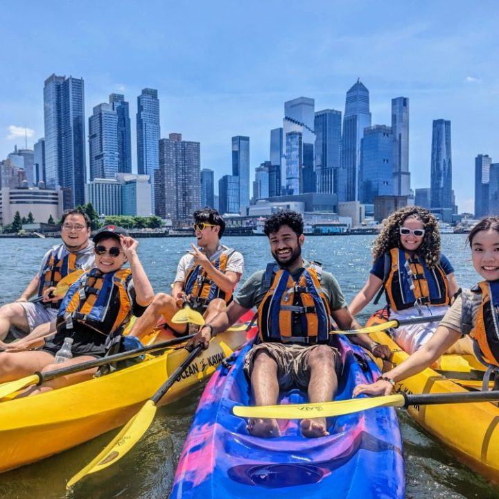 A small group of friends smiling and rafting their kayaks together on the Hudson River with a clear view of the Midtown Manhattan skyline.