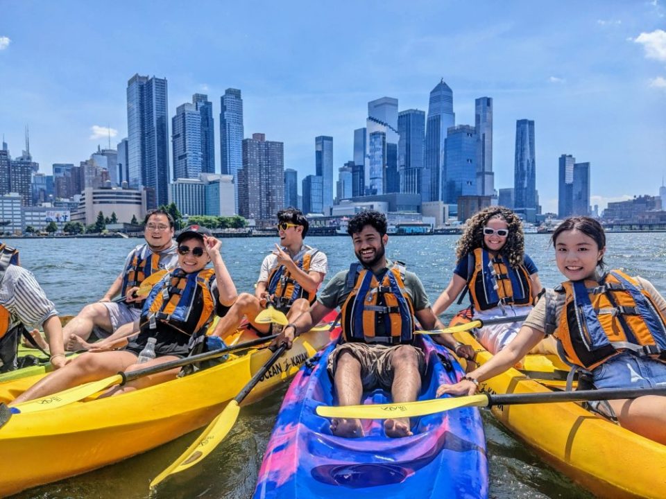 A small group of friends smiling and rafting their kayaks together on the Hudson River with a clear view of the Midtown Manhattan skyline.