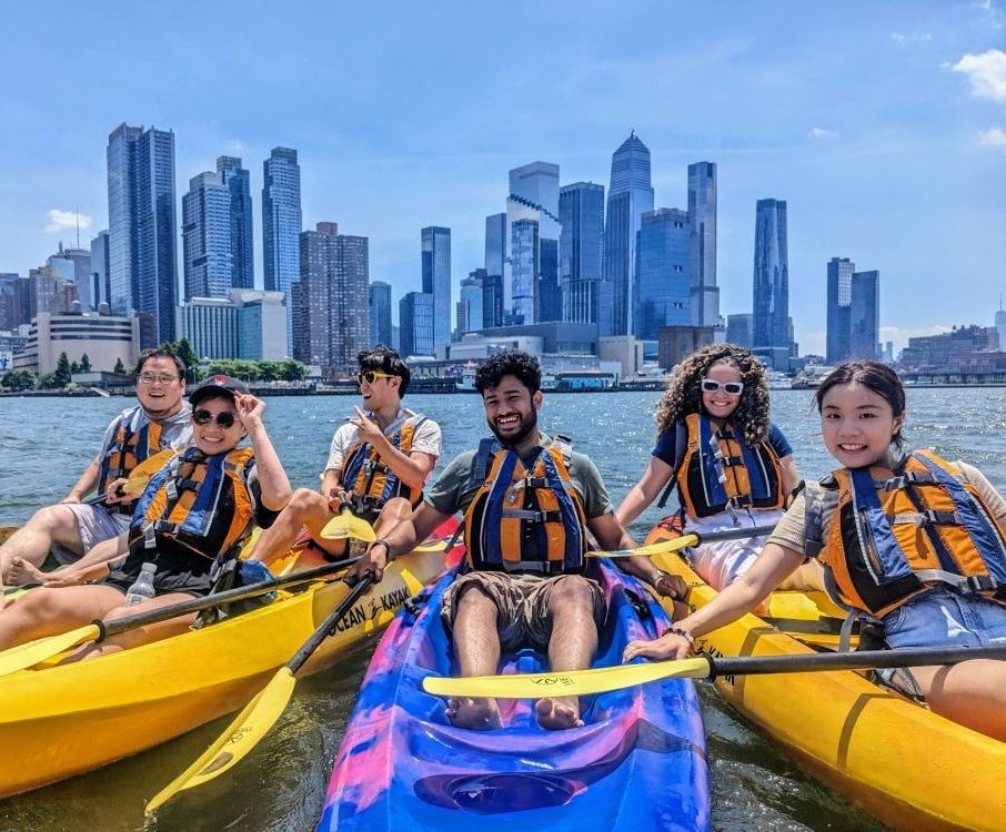 A small group of friends smiling and rafting their kayaks together on the Hudson River with a clear view of the Midtown Manhattan skyline.