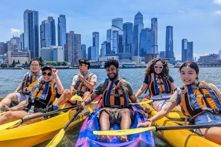 A small group of friends smiling and rafting their kayaks together on the Hudson River with a clear view of the Midtown Manhattan skyline.