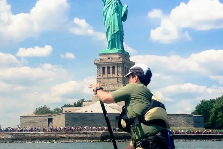 A paddleboarder on an advanced-level trip (denoted by a Single Black Diamond) riding in front of the Statue of Liberty on a sunny day with crowds of people standing on Liberty Island in the distance.