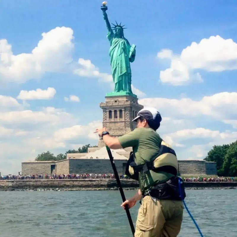 A paddleboarder on an advanced-level trip (denoted by a Single Black Diamond) riding in front of the Statue of Liberty on a sunny day with crowds of people standing on Liberty Island in the distance.